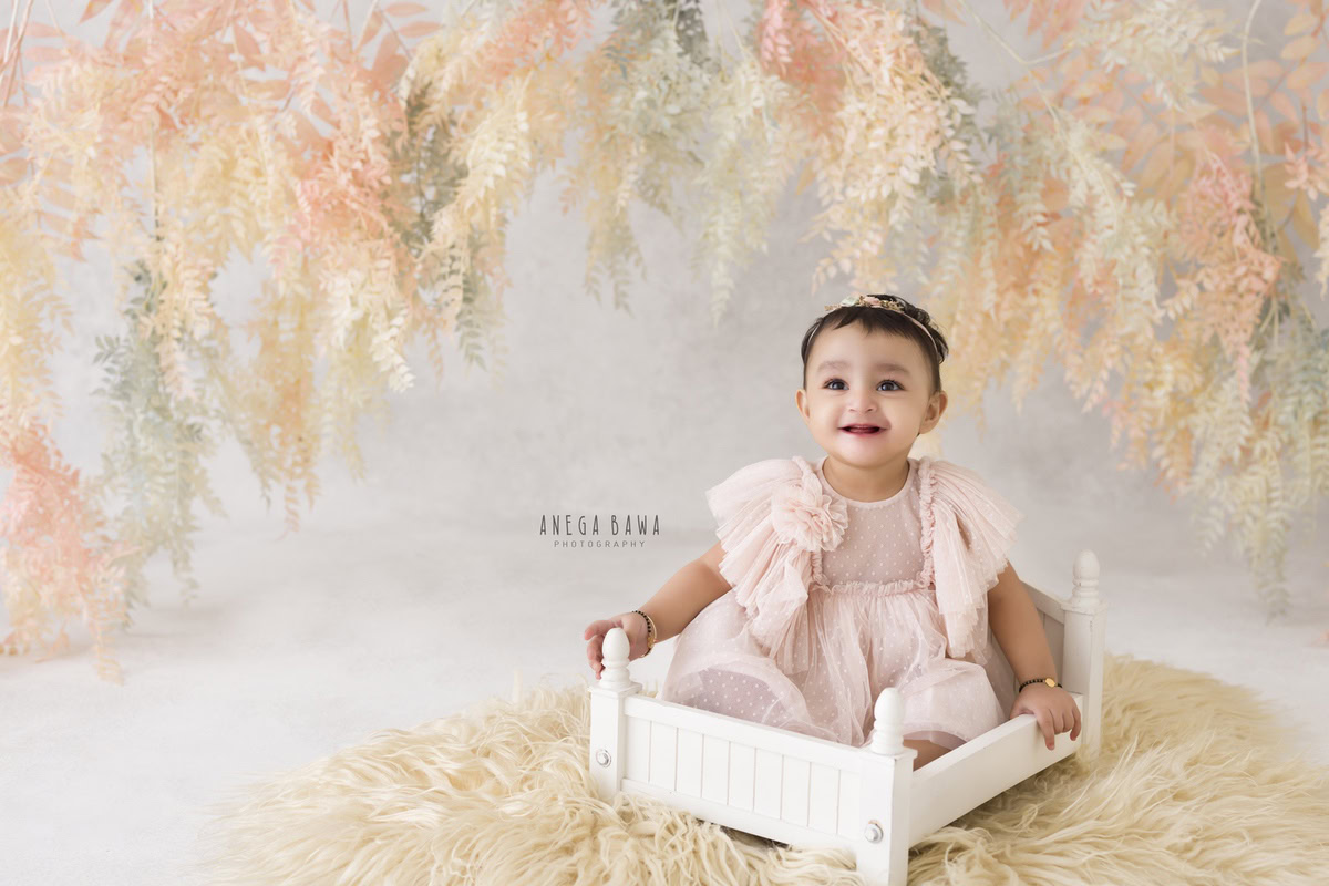 Girl seated in a pristine white cot on a cozy beige rug against a backdrop adorned with peachy leafy accents, marking her journey from 8 months to 1 year old during her first birthday photoshoot in Delhi by Anega Bawa, Gurgaon, Noida.