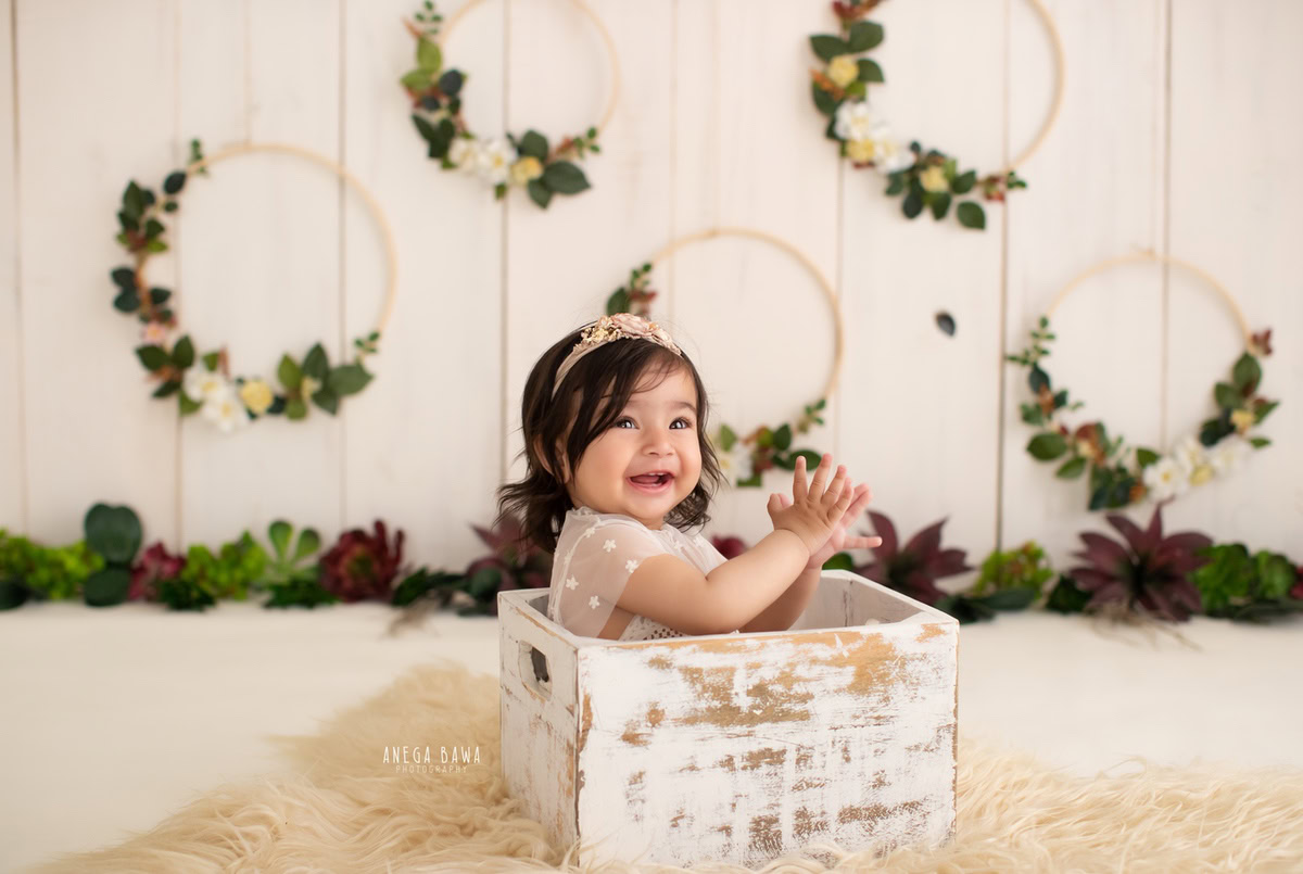Embrace the precious moments with your darling girl captured by Anega Bawa Photography in Delhi and Gurgaon. Seated in a cozy white cot amidst a serene beige backdrop, surrounded by wooden leafy frames, she shines in her innocence at 8 months to 1 year old.