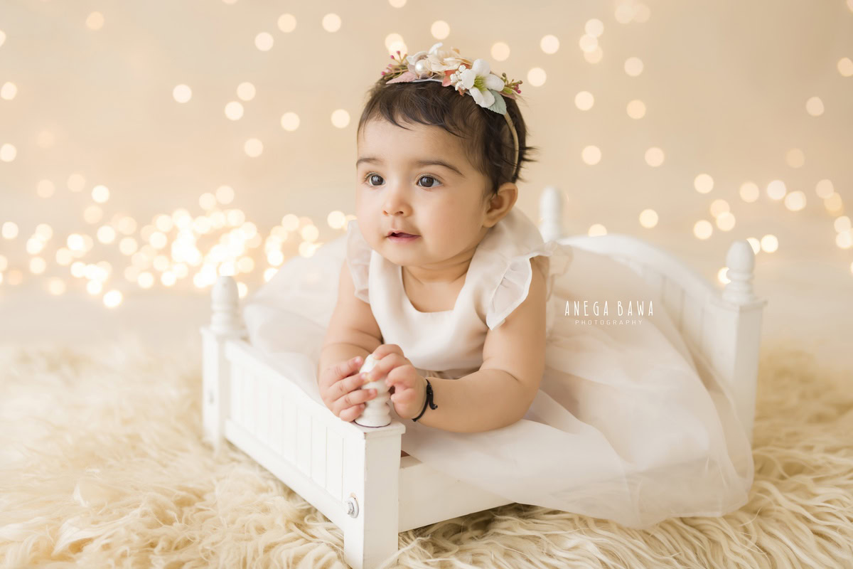 Behold your little princess seated gracefully in a pristine white cot, adorned with a cute headband that accentuates her innocence. With a cozy beige rug beneath her, and fairy lights illuminating the scene against a soothing beige backdrop, each snapshot captures her angelic charm. Experience the magic of sitter photography in Delhi and Gurgaon with Anega Bawa, where every moment is a treasure to cherish.