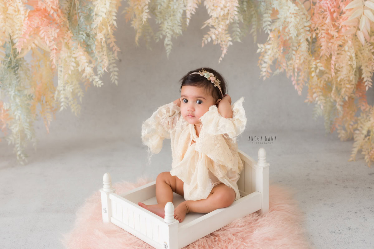 Girl seated in a pristine white cot wearing a cute headband, against a backdrop adorned with leafy accents, marking her journey from 8 months to 1 year old during her first birthday photoshoot in Delhi by Anega Bawa, Gurgaon, Noida.