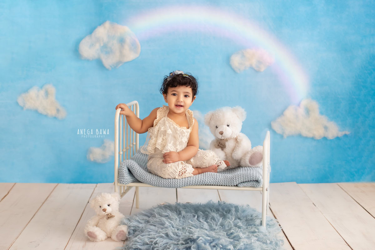 Captured by the talented lens of Anega Bawa in Delhi-Gurgaon, this delightful image features an adorable 8-month-old girl seated cozily in a white cot. Surrounding her are cuddly soft toys, adding a playful charm to the scene. The soft, comforting texture of the grey rug complements the serene ambiance created by the cloudy backdrop. With expert composition and attention to detail, this photograph beautifully captures the innocence and joy of childhood, making it a treasured memory for years to come.