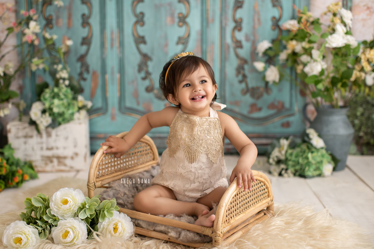 Delight in the charm of your little princess with Anega Bawa Photography in Delhi and Gurgaon. Seated gracefully in a wooden cot against a lush green backdrop, adorned with white flowers, she captures hearts at 8 months to 1 year old, creating treasured memories to cherish forever.