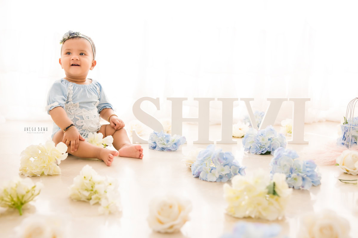 During a delightful sitter photoshoot in Delhi, Gurgaon by Anega Bawa, an adorable 8-month-old girl sits elegantly amidst a captivating scene. With a cute headband adorning her head, she poses beside a name frame placed on the floor, set against a serene white backdrop. This charming setup captures the innocence and charm of childhood, creating a timeless memory to cherish for years to come.