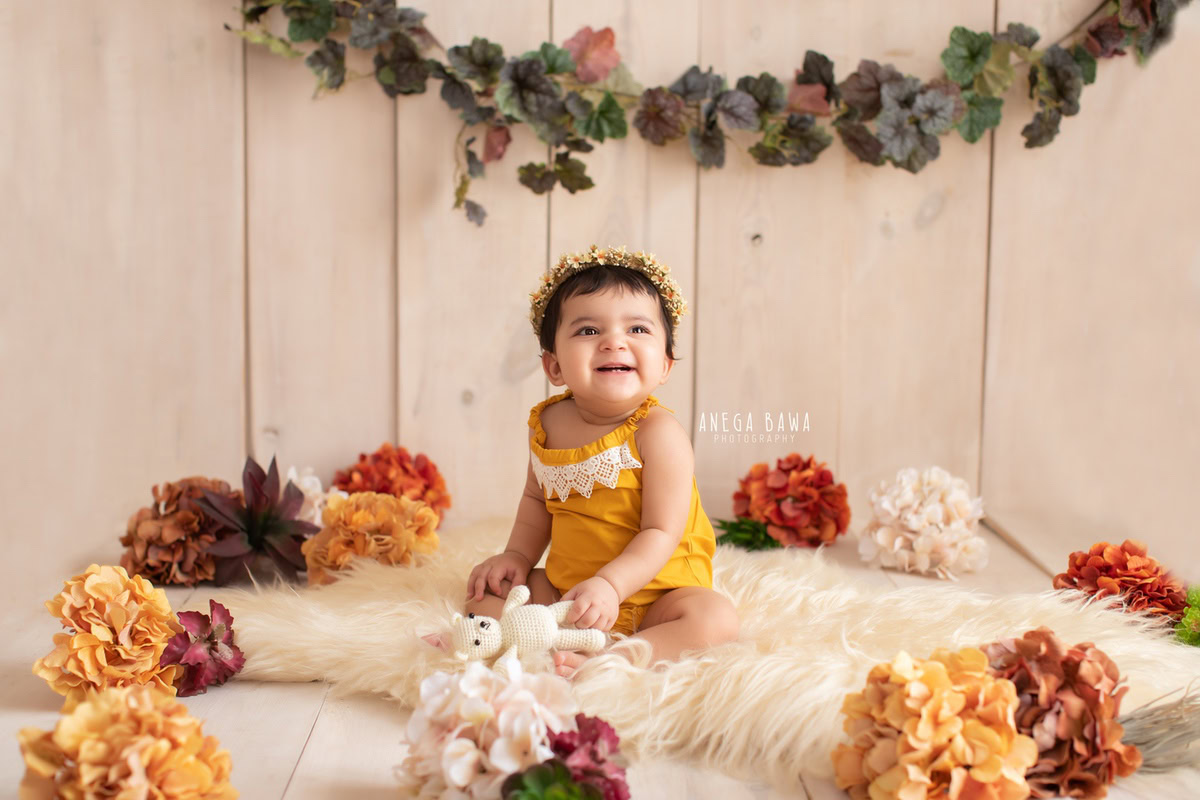 Captured by the talented Anega Bawa in Delhi-Gurgaon, this delightful image features an 8-month-old girl seated gracefully on a beige rug against a tranquil beige backdrop. Surrounding her are vibrant orange and brown flowers, accentuated by a delicate leafy fringe. The warm earthy tones evoke a sense of serenity, while the girl's serene expression adds to the charm of the scene. Through expert composition and artistry, this photograph beautifully encapsulates the innocence and beauty of infancy, making it a cherished keepsake for the family.