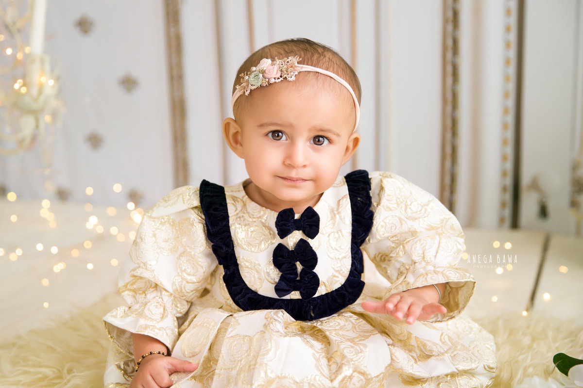 In a delightful sitter photoshoot by Anega Bawa in Delhi, Gurgaon, a precious 8-month-old girl sits gracefully on a beige rug. Fairy lights twinkle softly around her, casting a magical glow against the pristine white backdrop. This enchanting scene captures the innocence and wonder of childhood, creating cherished memories for years to come.