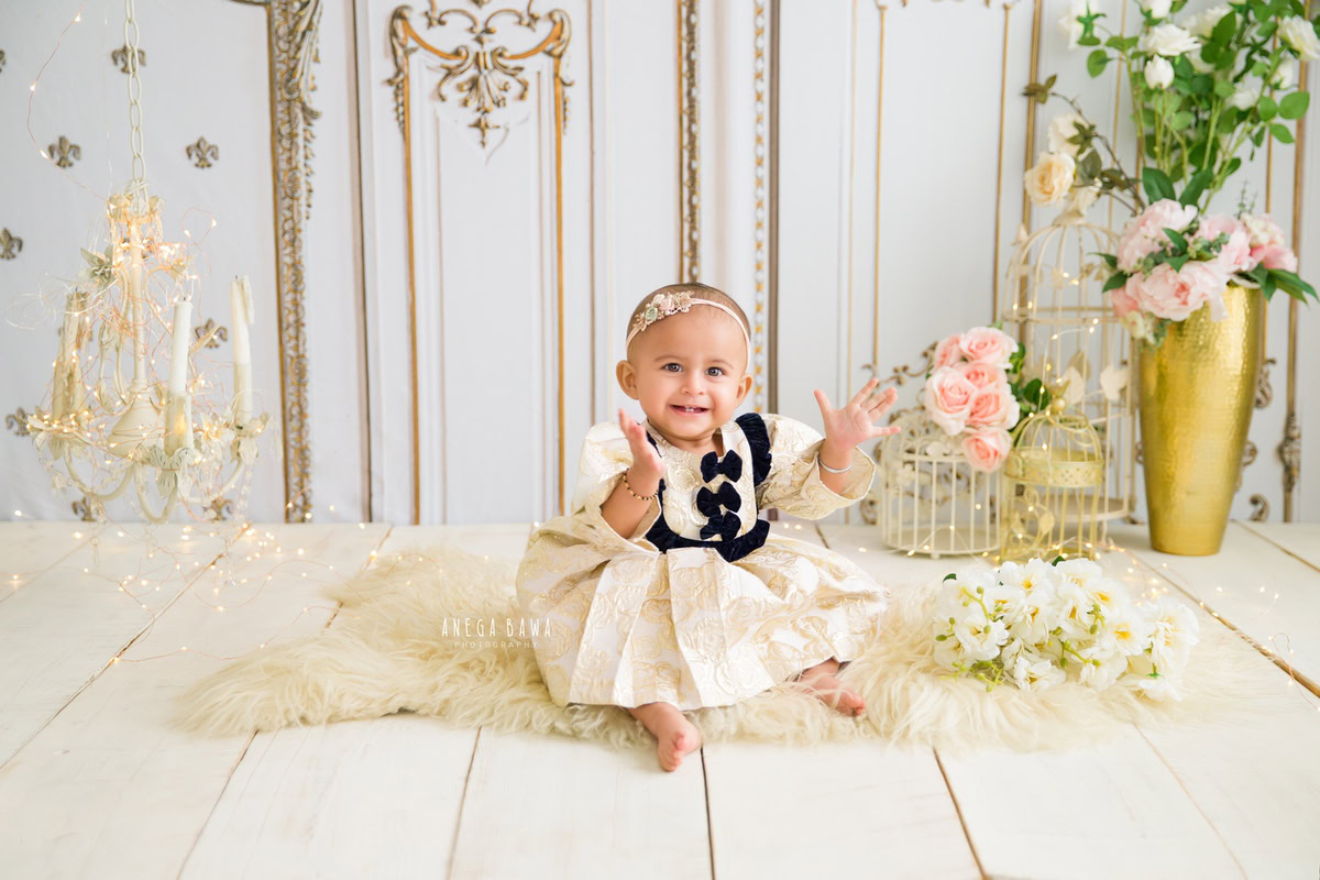 Captured during a heartwarming sitter photoshoot by Anega Bawa in Delhi, Gurgaon, is an adorable 8-month-old girl seated elegantly on a beige rug. Beside her, a golden flower vase and a candle stand add a touch of sophistication, while white castles create a dreamy backdrop. This enchanting scene captures the innocence and beauty of childhood, preserving precious moments for a lifetime.