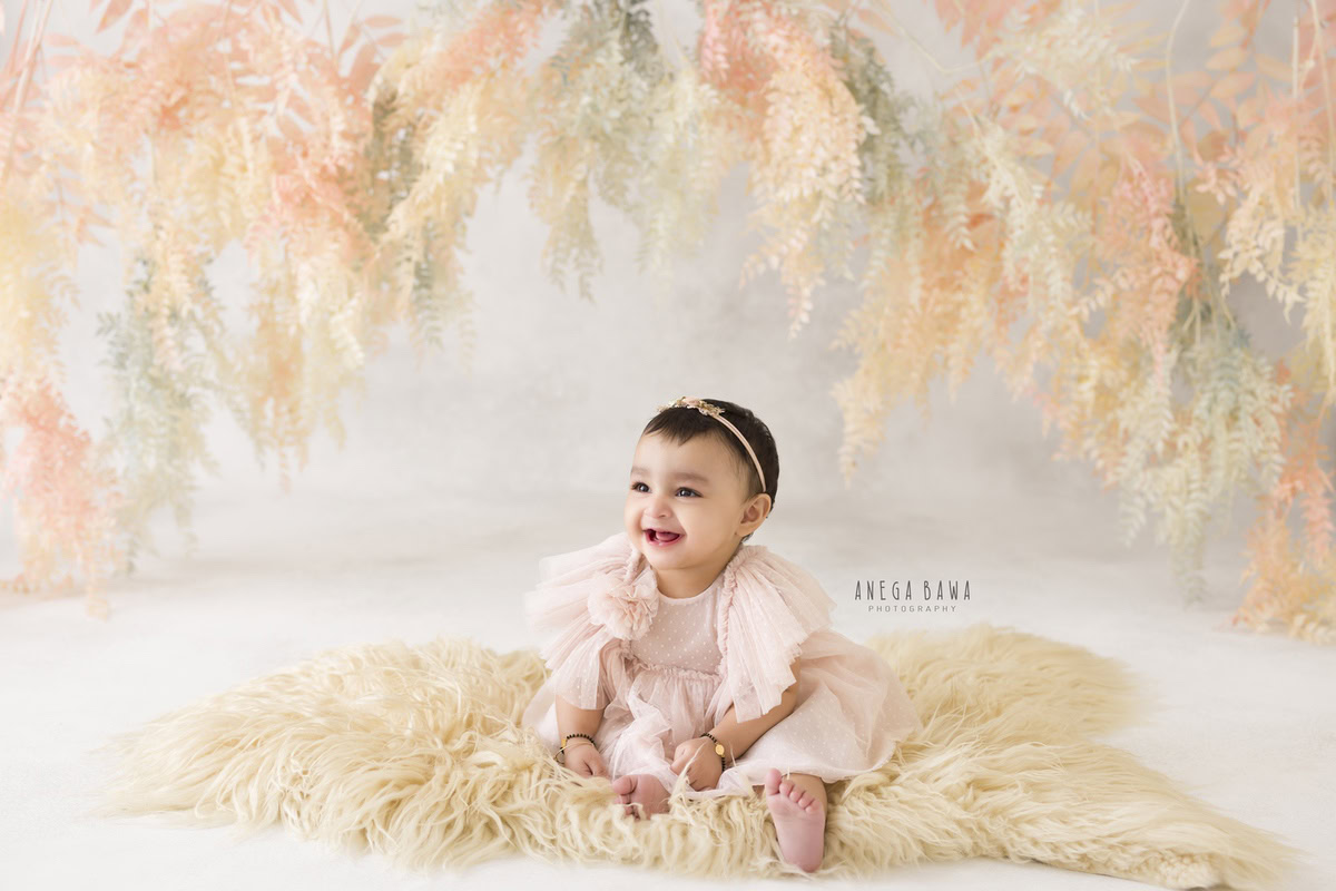 Girl seated on a cozy beige rug against a backdrop adorned with leafy peachy accents, marking her journey from 8 months to 1 year old during her first birthday photoshoot in Delhi by Anega Bawa, Gurgaon, Noida.