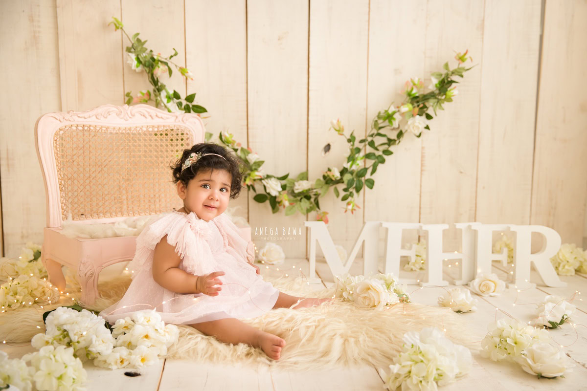 In this delightful moment captured by Anega Bawa's sitter photography in Delhi, Gurgaon, a charming 8-month-old girl sits on a beige rug. A name frame rests on the floor beside her, surrounded by white flowers. The scene is adorned with a leafy fringe on the wall, creating a picturesque backdrop that adds to the magic of the moment.