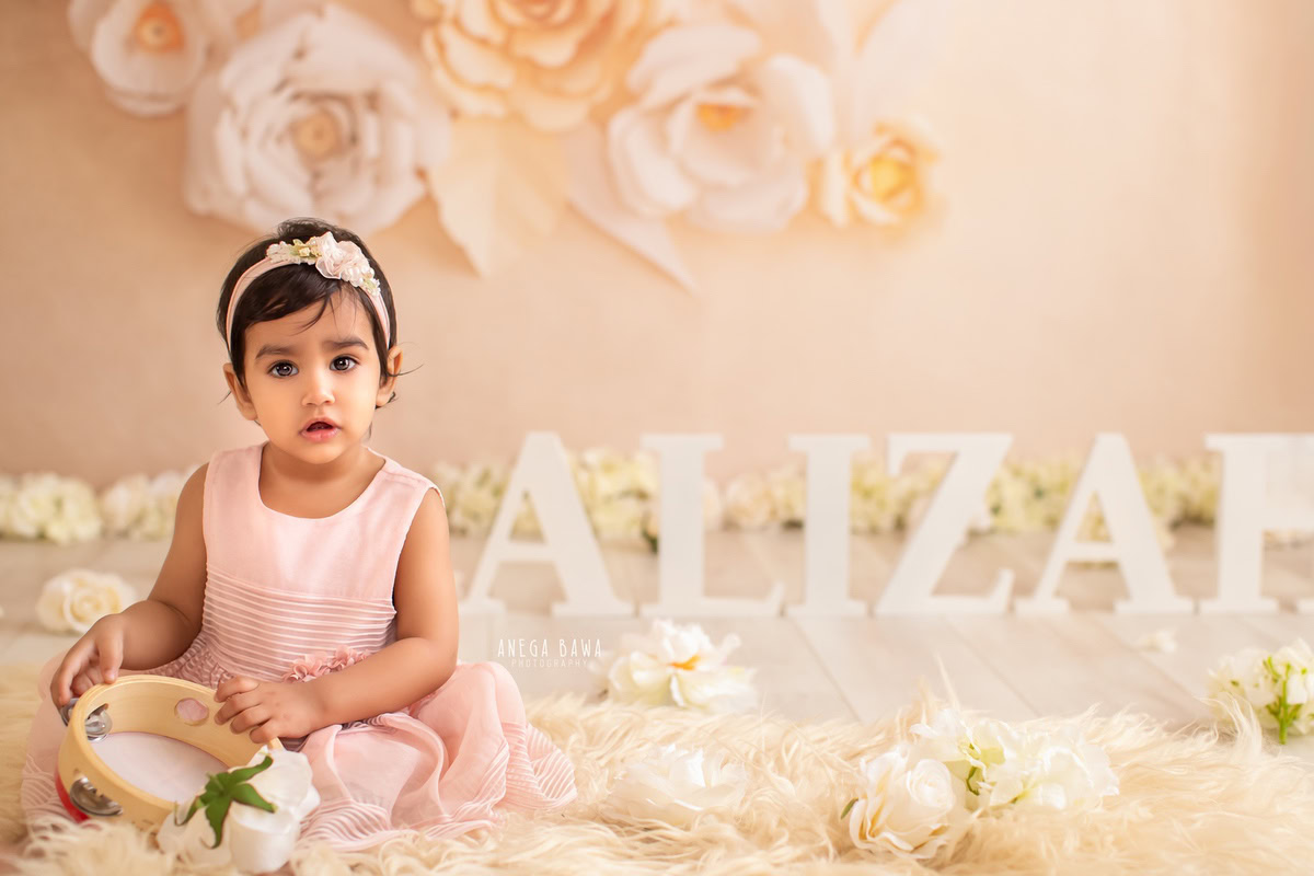 Captured by Anega Bawa's sitter photography in Delhi, Gurgaon, this adorable scene features an 8-month-old girl seated on a beige rug amidst a backdrop of vibrant floral patterns. A name frame rests on the floor beside her, adding a personal touch to the charming setting.