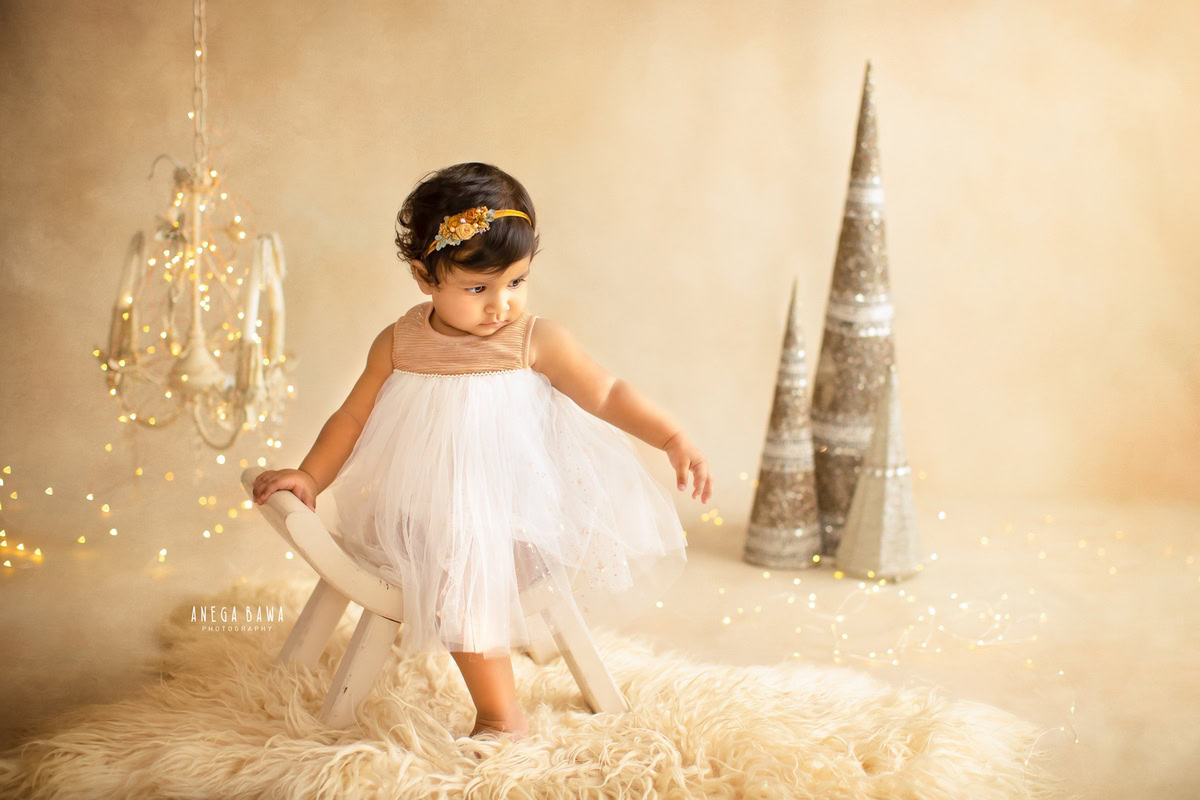 In a captivating setup against a beige backdrop, a 7-month-old girl sits gracefully on a curvy chair, surrounded by the warm glow of golden lights emanating from candlestands. This enchanting scene captures the essence of her first year in a baby pre-birthday photoshoot in Delhi, Anega Bawa, Gurgaon, and Noida.