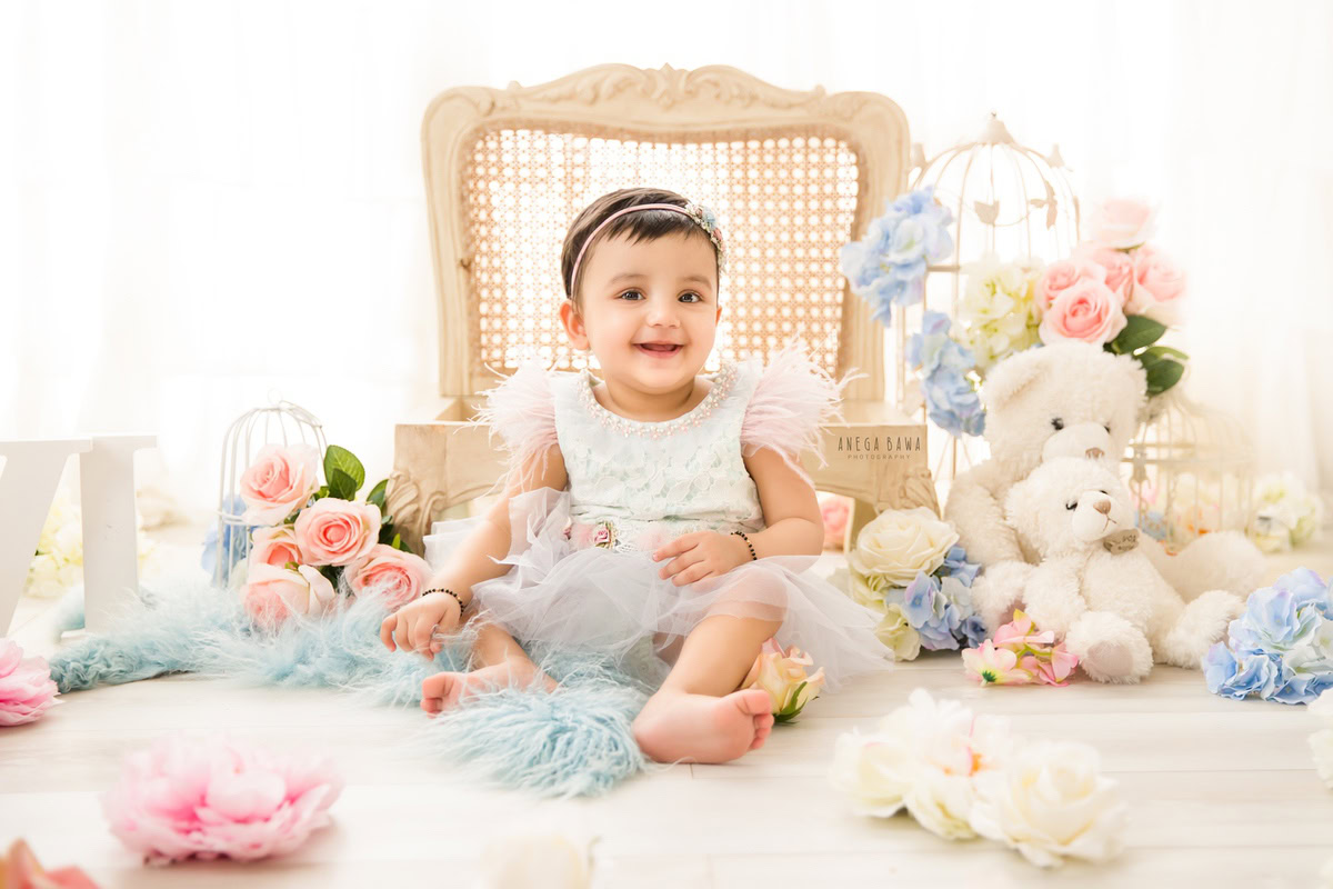 Capturing the precious moments of your little princess, Anega Bawa Photography in Delhi and Gurgaon invites you to witness her seated on a wooden chair amidst blue and pink flowers. With a soft toy by her side against a serene white backdrop, she exudes innocence and charm at 7 months to 1 year old.