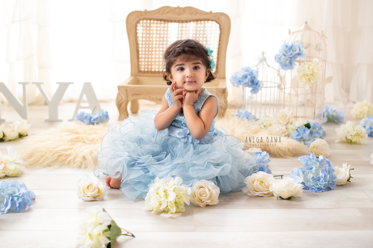 In a delightful scene by Anega Bawa Photography in Delhi and Gurgaon, your little princess is seated on a beige rug surrounded by blue and white flowers. With white castles and a wooden chair adding to the charm, she radiates innocence and joy at 7 months to 1 year old.