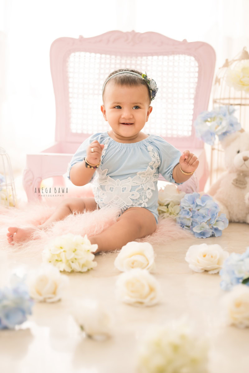 In this delightful image from Anega Bawa's sitter photography sessions in Delhi, Gurgaon, a girl, aged 7 months to 1 year old, is captured seated on the floor against a serene white backdrop adorned with blue and white flowers.