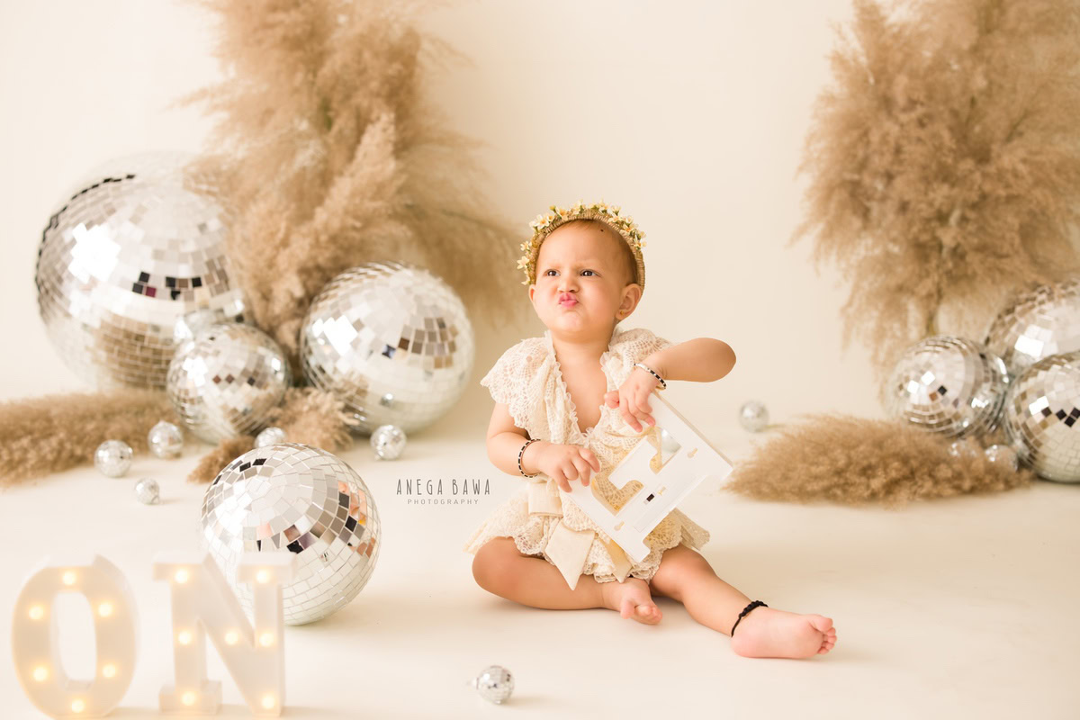 Captured during Anega Bawa's sitter photography sessions in Delhi, Gurgaon, this charming image features a girl, aged 7 months to 1 year old, striking a pouty pose while seated on the floor. She wears a tiara band, adding a touch of elegance to the scene, against a beige backdrop adorned with delicate discoballs.