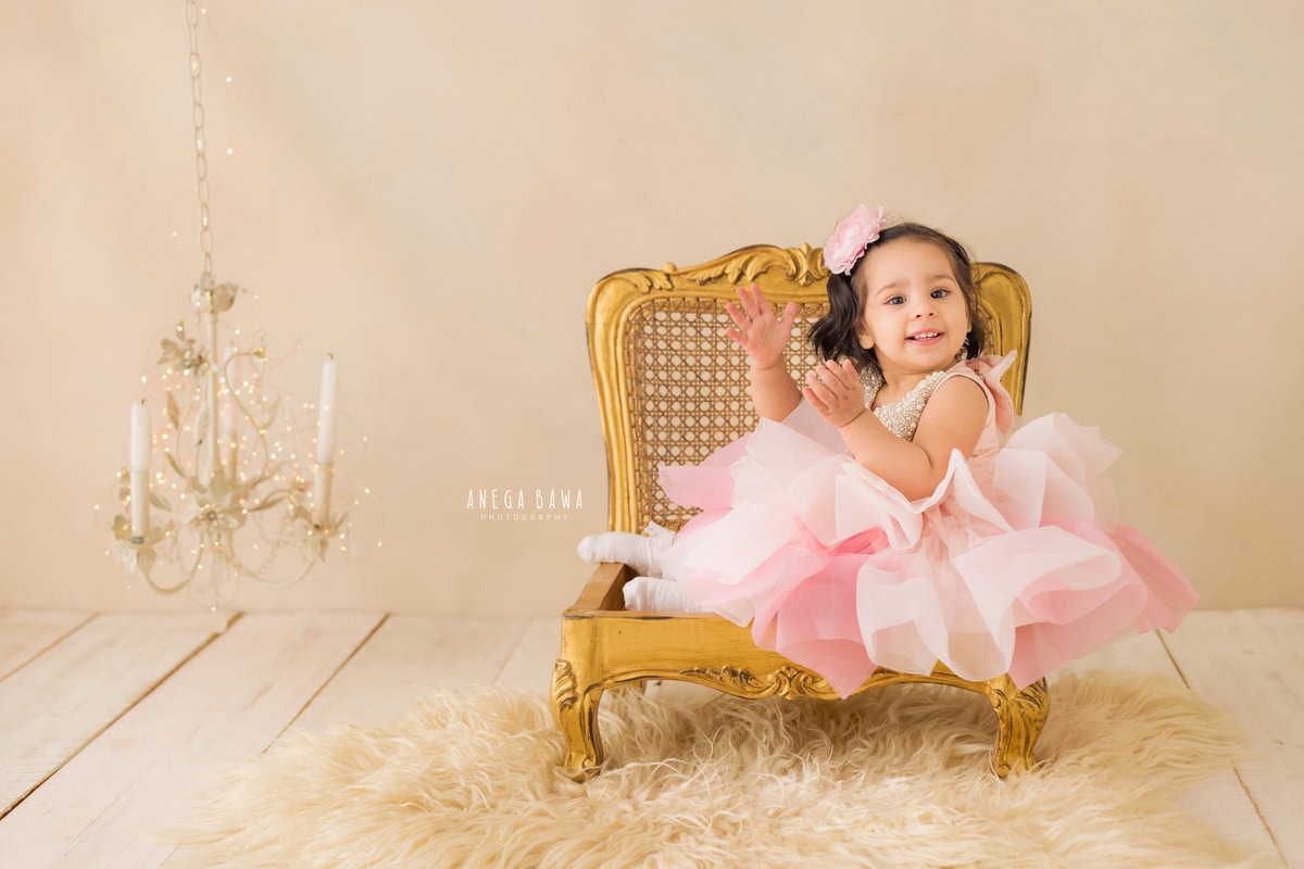 Girl seated on a regal golden chair atop a cozy beige rug against a serene beige backdrop, with a candle stand nearby, capturing her journey from 7 months to 1 year old during her first birthday photoshoot in Delhi by Anega Bawa, Gurgaon, Noida.