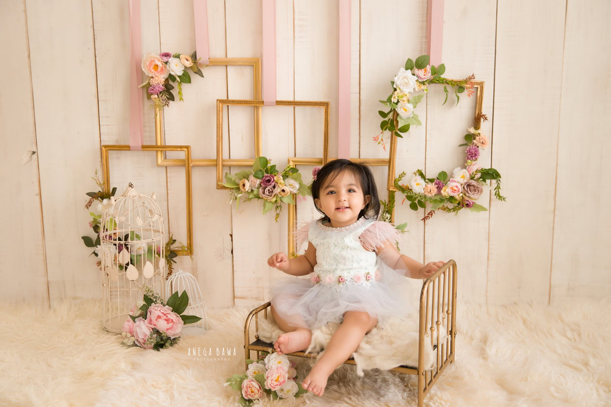 In a serene setting captured by Anega Bawa Photography in Delhi and Gurgaon, your little princess sits on a golden cot amidst floral frames against a calming beige backdrop. With delicate pink flowers enhancing the scene, she exudes pure charm and innocence at 7 months to 1 year old.