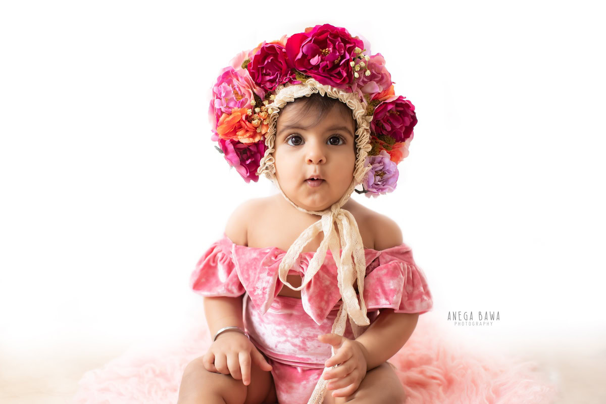 In this delightful photo by Anega Bawa, taken in Gurgaon-Delhi, a 7-month-old girl sits gracefully on a soft pink rug against a pristine white backdrop. Adorned with a charming pink floral head cap, she exudes innocence and sweetness, capturing the essence of childhood. The expertly captured moment showcases the tender beauty of infancy, making it a precious memory for the family to cherish.
