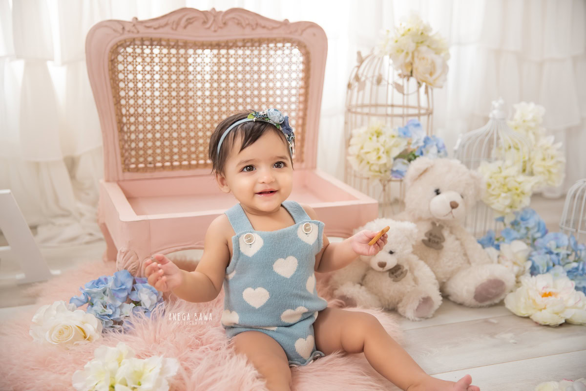 In a delightful scene painted by Anega Bawa Photography in Delhi and Gurgaon, your little princess sits on a pink rug surrounded by teddy bears, blue and white flowers, and castles against a pristine white backdrop. At 7 months to 1 year old, she radiates joy and innocence, creating enchanting memories to cherish.