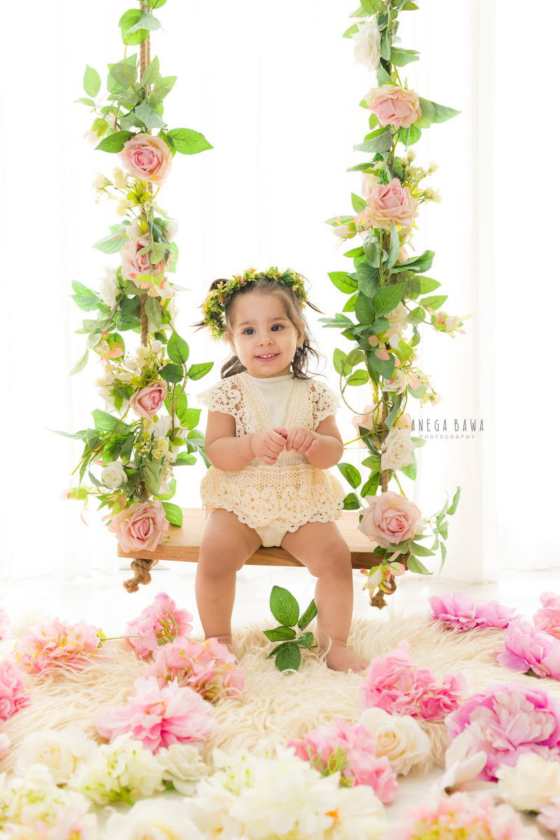 Girl seated on a charming swing surrounded by pink and white flowers on the floor against a pristine white backdrop, marking her journey from 7 months to 1 year old during her first birthday photoshoot in Delhi by Anega Bawa, Gurgaon, Noida.