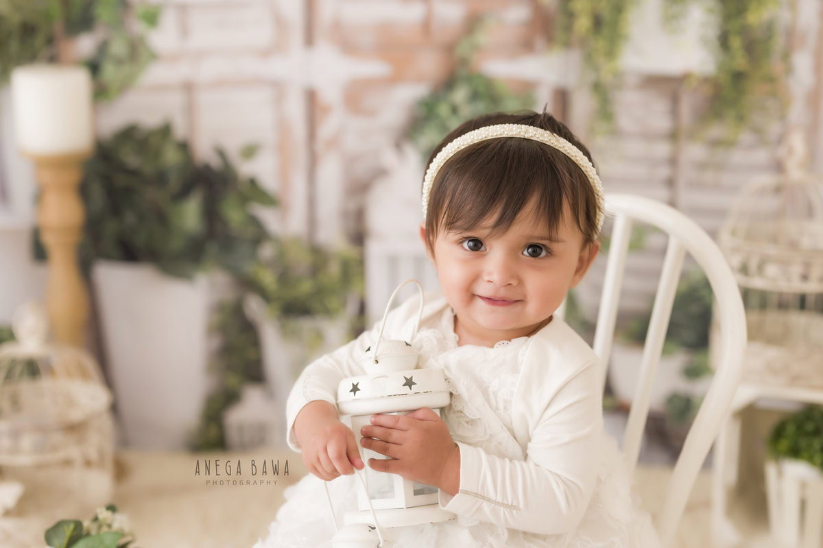 In a timeless setting crafted by Anega Bawa Photography, your 13-month to 1-year-old princess sits gracefully on a pristine white chair. Illuminated by the soft glow of candlelight and surrounded by verdant leafy pots, she enchants against a pure white backdrop. Capture these moments of elegance and purity in Delhi and Gurgaon with Anega Bawa Photography.