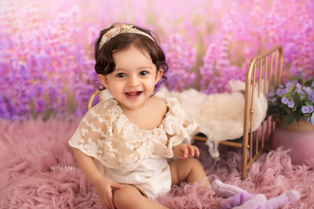 In this enchanting photo by Anega Bawa, taken in Gurgaon-Delhi, a 13-month-old girl sits gracefully on a soft pink rug against a serene lavender backdrop. Adorned with a cute headband, she exudes innocence and charm, capturing the essence of childhood. The expertly captured moment showcases the tender beauty of infancy, making it a precious memory for the family to cherish.