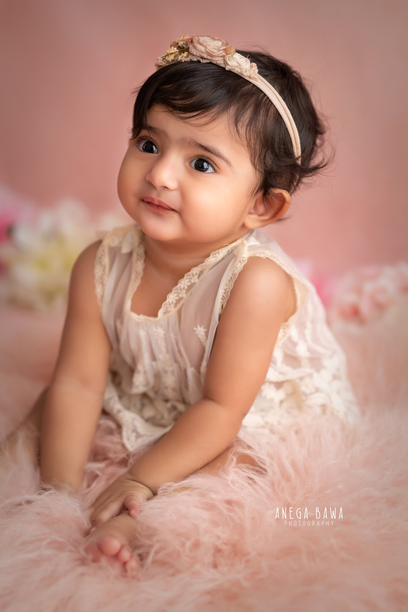 Capturing the innocence and charm of a 13-month-old in Gurgaon, Anega Bawa, Delhi, this photo features a girl seated on a pink rug against a matching pink backdrop. With delicate details and a soft palette, the image beautifully portrays the sweetness of this little sitter at this precious age.