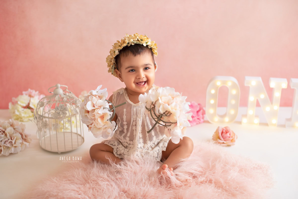 In a picturesque setting crafted by Anega Bawa Photography in Delhi and Gurgaon, your adorable princess sits on a pink rug adorned with white flowers against a charming pink backdrop. At 13 months to 1 year old, she exudes grace and sweetness, capturing timeless memories to treasure forever.
