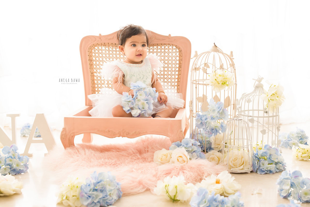 In this enchanting capture from Anega Bawa's sitter photography sessions in Delhi, Gurgaon, a girl aged 13 months to 1 year old sits gracefully on a pink wooden chair. The scene is adorned with white castles, a pink rug, and a pristine white backdrop, creating a whimsical ambiance for the delightful moment.