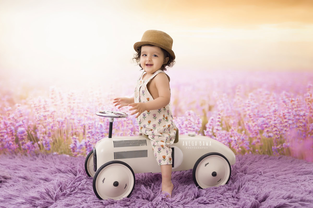 Immerse in the delightful sight of your 13-month to 1-year-old princess seated on a toy scooter, adorned with a sweet hat. Against a backdrop reminiscent of soothing lavender hues, set on a lavender rug, she radiates charm and innocence. Preserve these precious moments with Anega Bawa Photography, offering enchanting photo sessions in Delhi and Gurgaon.