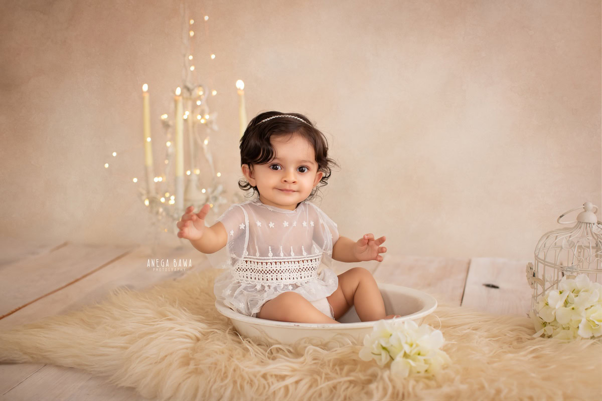 In a delightful scene curated by Anega Bawa Photography in Delhi and Gurgaon, your little princess sits in a tub on a cozy beige rug against a serene beige backdrop, surrounded by flickering candles and warm lights. At 13 months to 1 year old, she radiates innocence and charm, creating timeless memories to cherish forever.