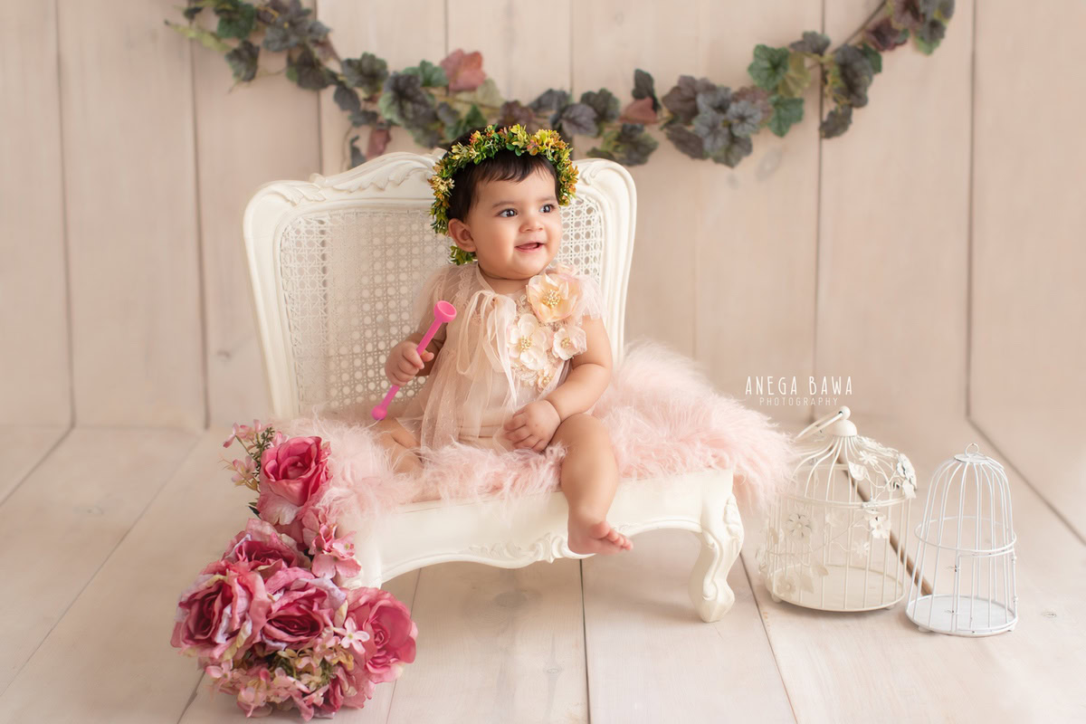Girl seated on a white chair amidst white castles with a floral fringe on the wall against a serene beige backdrop, marking her journey from 13 months to 1 year old during her first birthday photoshoot in Delhi by Anega Bawa, Gurgaon, Noida.