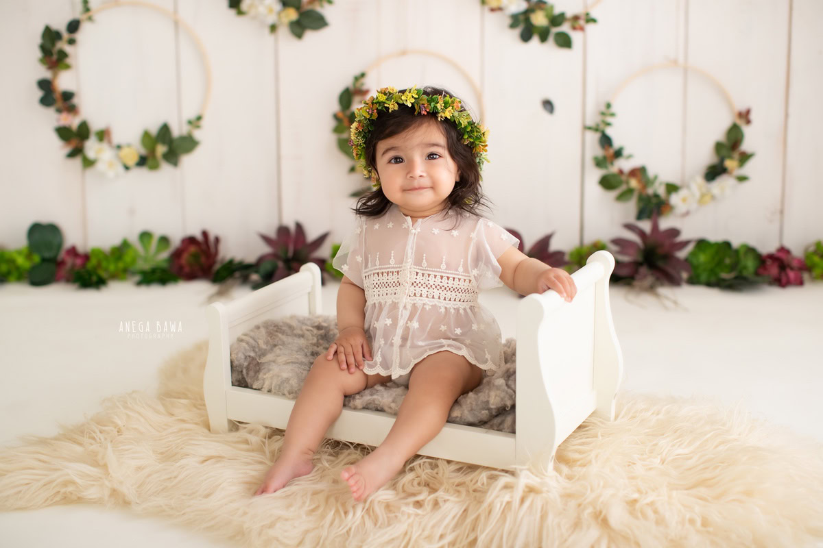 Girl seated in a pristine white cot against a serene beige backdrop, with a cozy beige rug and a leafy wooden frame on the wall, marking her journey from 13 months to 1 year old during her first birthday photoshoot in Delhi by Anega Bawa, Gurgaon, Noida.