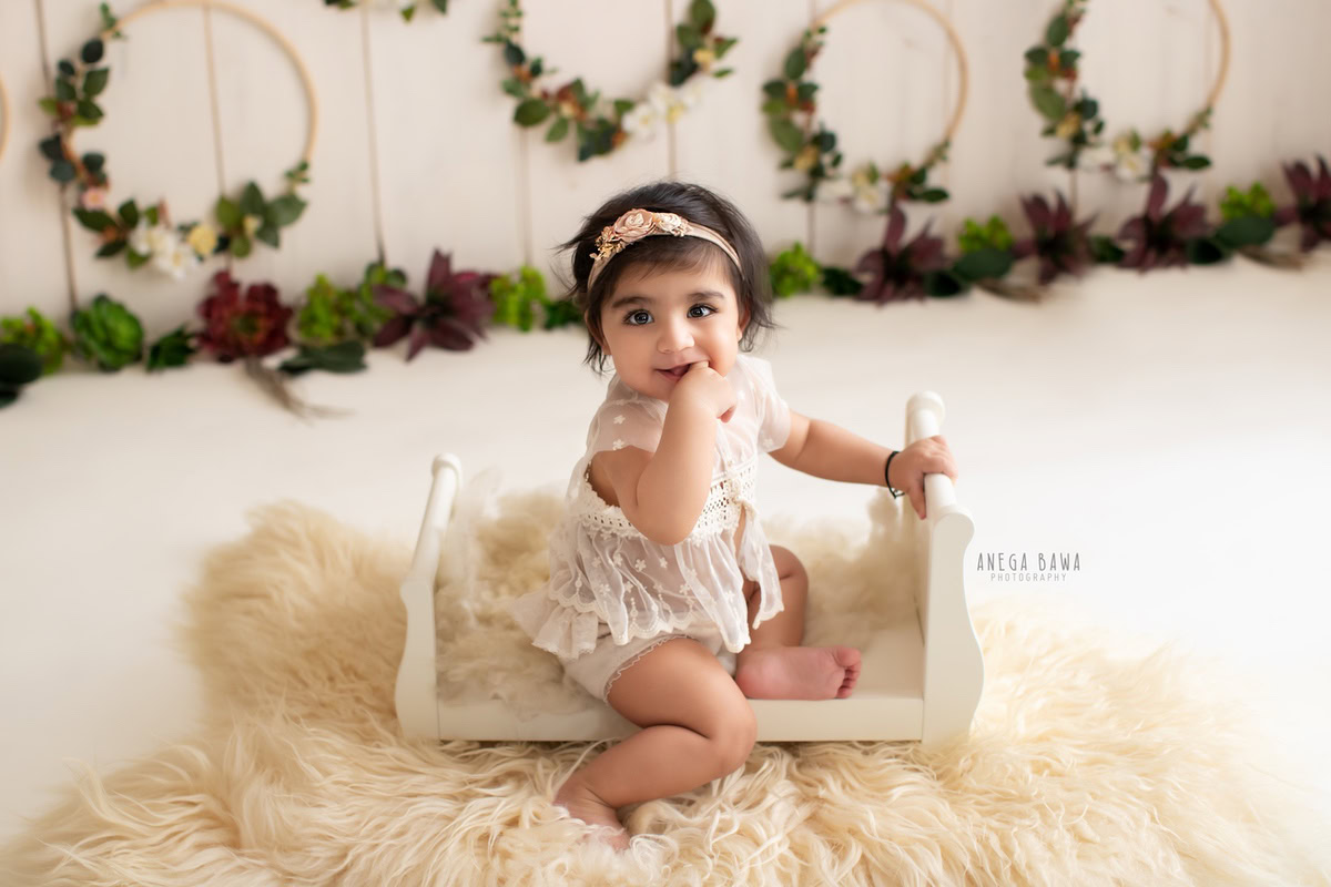 Girl seated in a pristine white cot on a cozy beige rug surrounded by floral wooden frames, marking her journey from 13 months to 1 year old during her first birthday photoshoot in Delhi by Anega Bawa, Gurgaon, Noida.