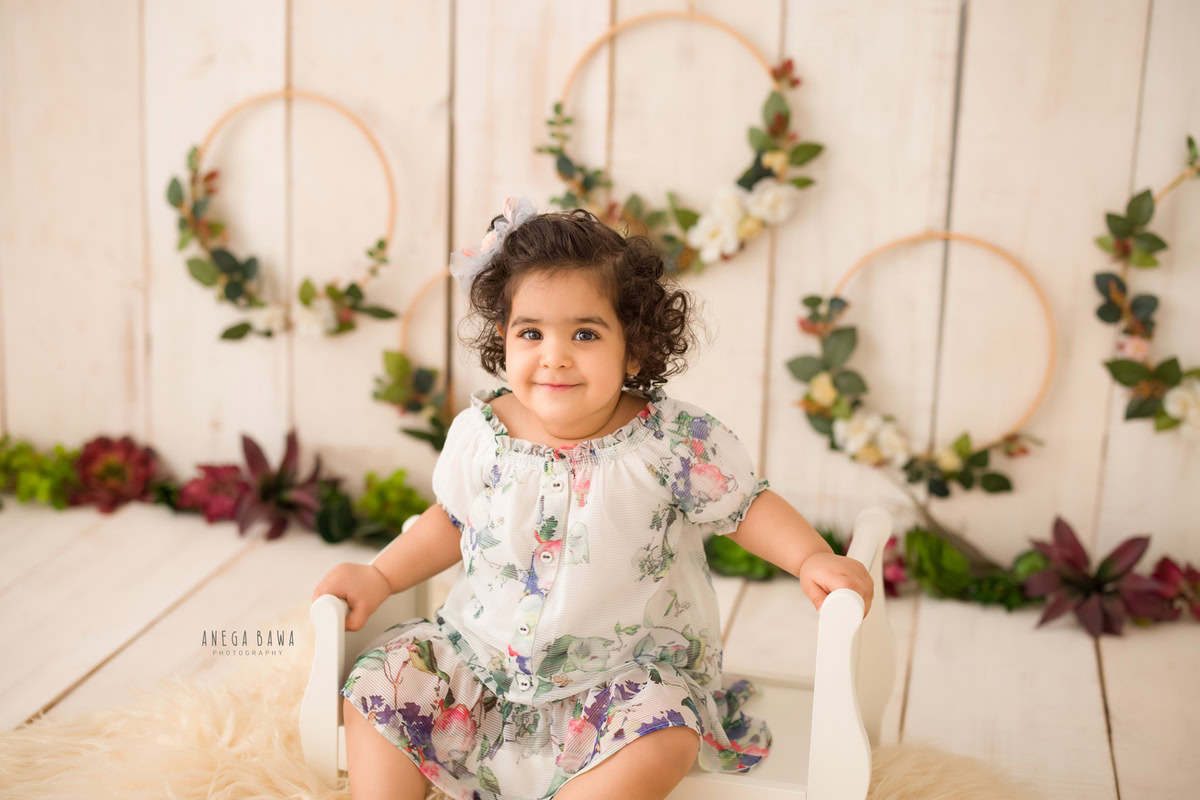 Captured during Anega Bawa's sitter photoshoot in Delhi, Gurgaon, this adorable image features a girl aged 13 months to 1 year old seated on a white cot. The backdrop showcases charming floral wooden frames against a soothing beige backdrop, adding a touch of elegance to the scene.