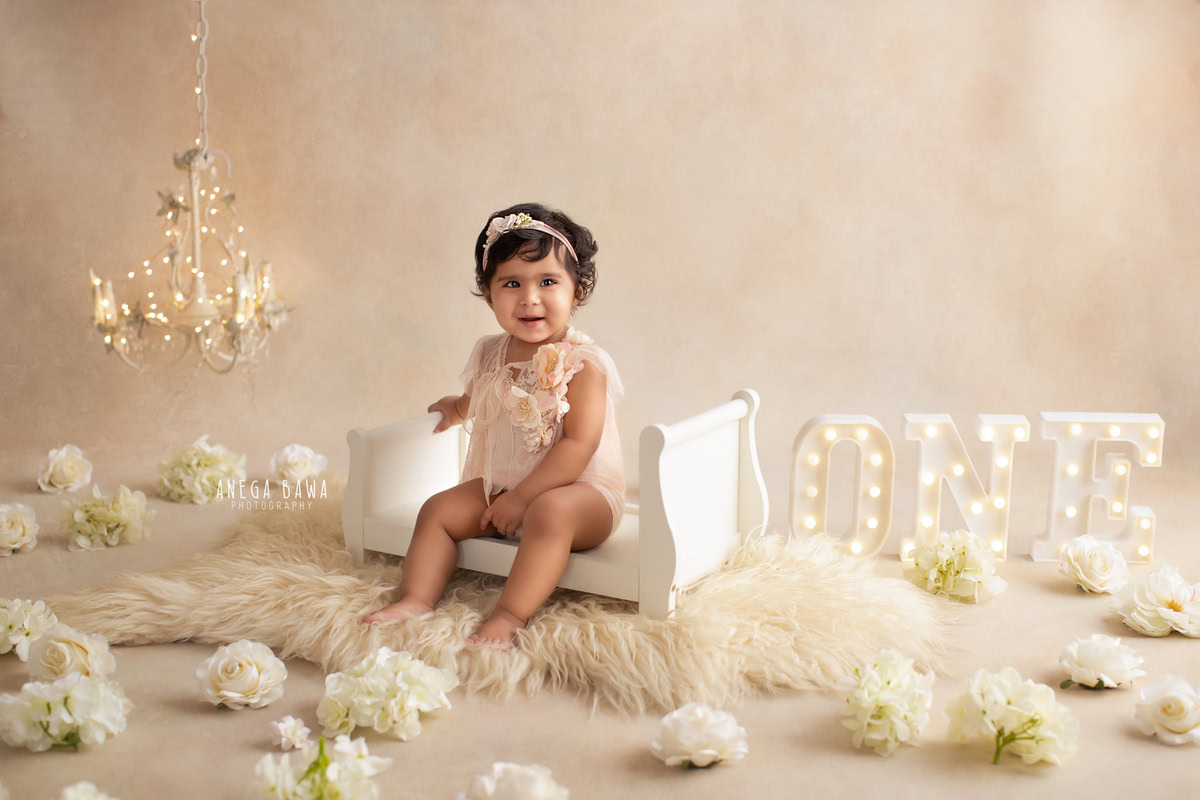 In a serene setting by Anega Bawa Photography in Delhi and Gurgaon, your little princess, aged 13 months to 1 year, sits gracefully on a pristine white cot surrounded by delicate white flowers. The soft glow of candle stands with lights adds a touch of warmth to the beige backdrop, capturing her innocence and charm in timeless frames.