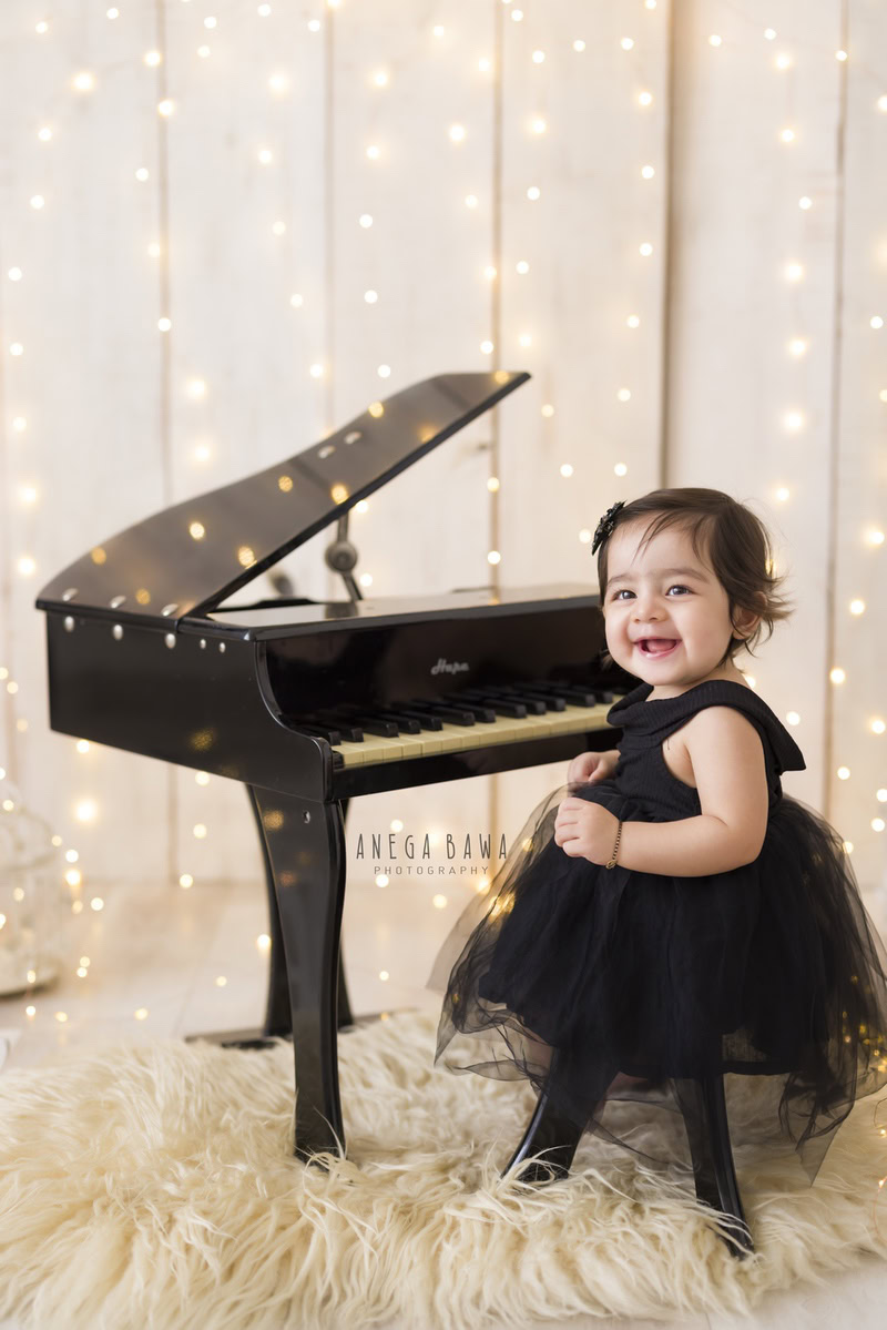 In this adorable photo from Gurgaon, Anega Bawa, Delhi, a 13-month-old girl is seated next to a piano, surrounded by a beige rug and twinkling fairy lights against a matching backdrop. The scene captures the innocence and curiosity of the little sitter, creating a charming moment to treasure.