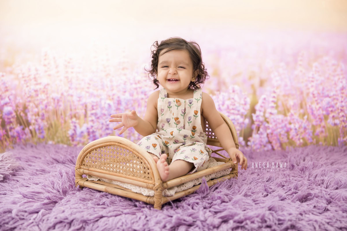 Capture the grace and innocence of your 13-month to 1-year-old princess as she sits on a wooden brown cot, surrounded by the tranquility of a lavender backdrop. Set on a soft lavender rug, every detail is accentuated, creating timeless memories to cherish. Book your photoshoot with Anega Bawa Photography in Delhi and Gurgaon to preserve these precious moments forever.