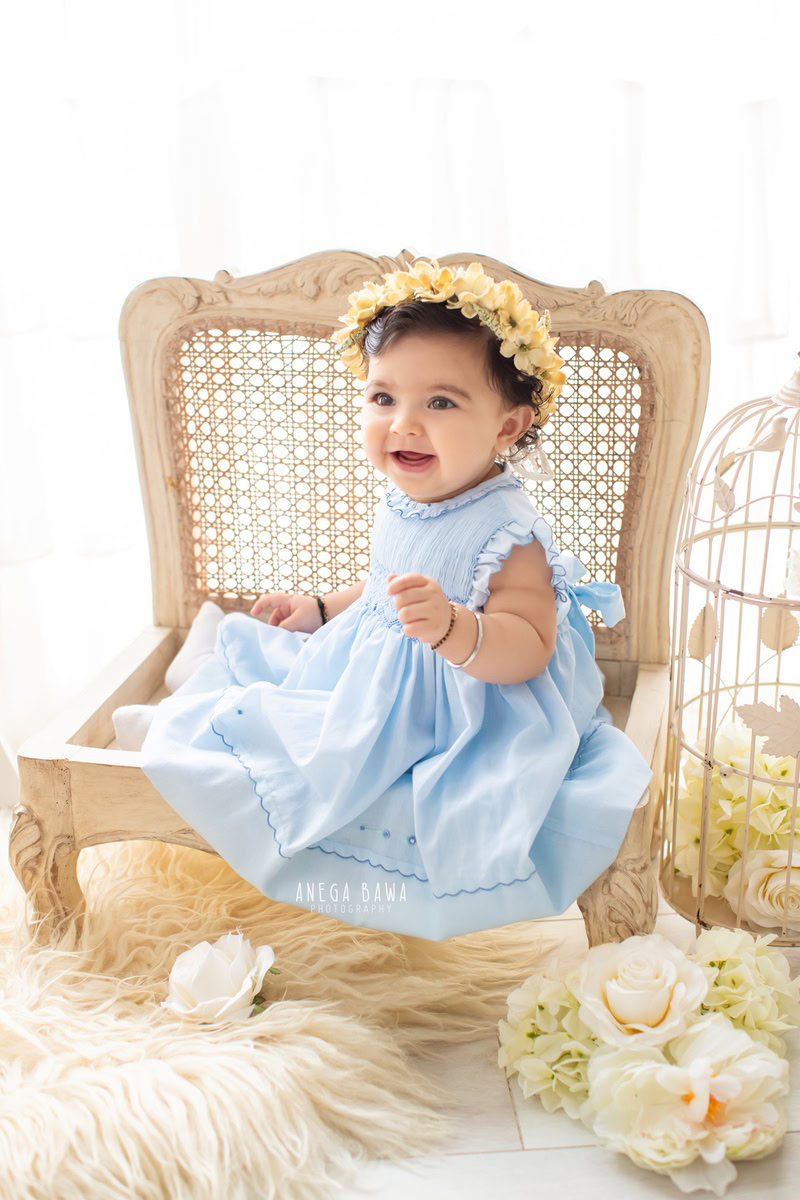 Girl seated on a wooden chair with a tiara band against a pristine white backdrop, accompanied by a cozy beige rug, commemorating her journey from 13 months to 1 year old during her first birthday photoshoot in Delhi by Anega Bawa, Gurgaon, Noida.