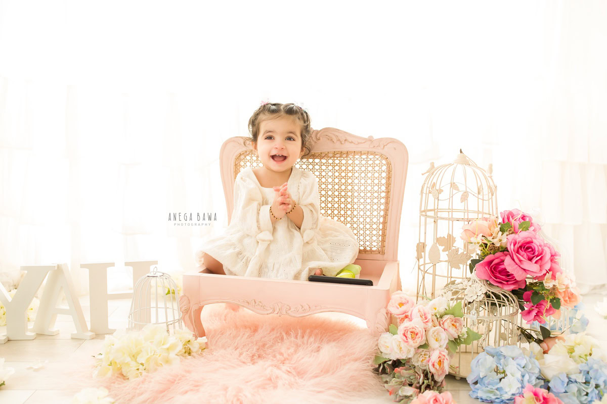 In a delightful photoshoot by Anega Bawa Photography in Delhi and Gurgaon, your 13-month to 1-year-old princess sits gracefully on a wooden chair against a pristine white backdrop. Adorned with multicolored flowers and surrounded by a pink rug, she radiates joy and innocence, creating precious memories to cherish forever.