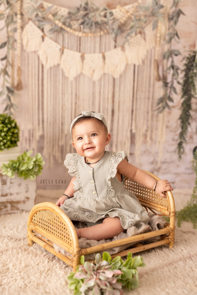 In this delightful capture from Anega Bawa's sitter photoshoot in Delhi, Gurgaon, a charming girl, aged 13 months to 1 year old, sits gracefully on a wooden cot. The scene is adorned with leafy bunches on the floor and fringes adorning the wall, all set against a serene beige backdrop.