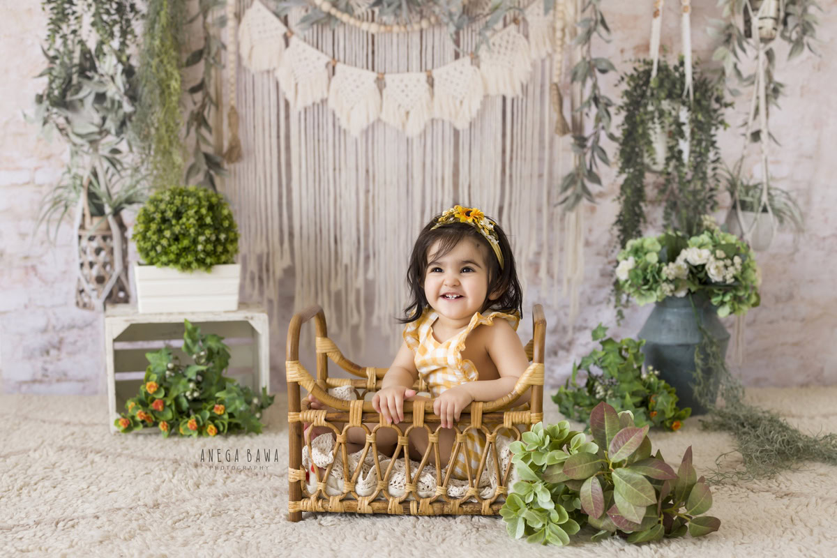 Immerse in the serenity of your 13-month to 1-year-old girl seated in a wooden cot, adorned with delicate blue pots against a pristine white backdrop. This charming setup, captured by Anega Bawa Photography in Delhi and Gurgaon, ensures that every moment is etched in timeless elegance. Book your session now to preserve these precious memories forever.