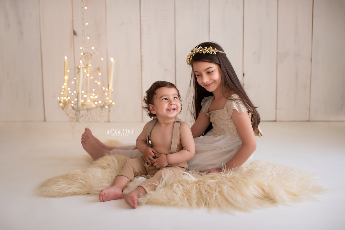 In the warm glow of golden lights at Anega Bawa Photography's studio in Delhi and Gurgaon, your 14-month to 1-year-old princess and her sibling share a tender moment on a cozy beige rug against a neutral beige backdrop. The flickering candlestand adds a touch of magic to the scene, creating a timeless photograph capturing their sibling bond.