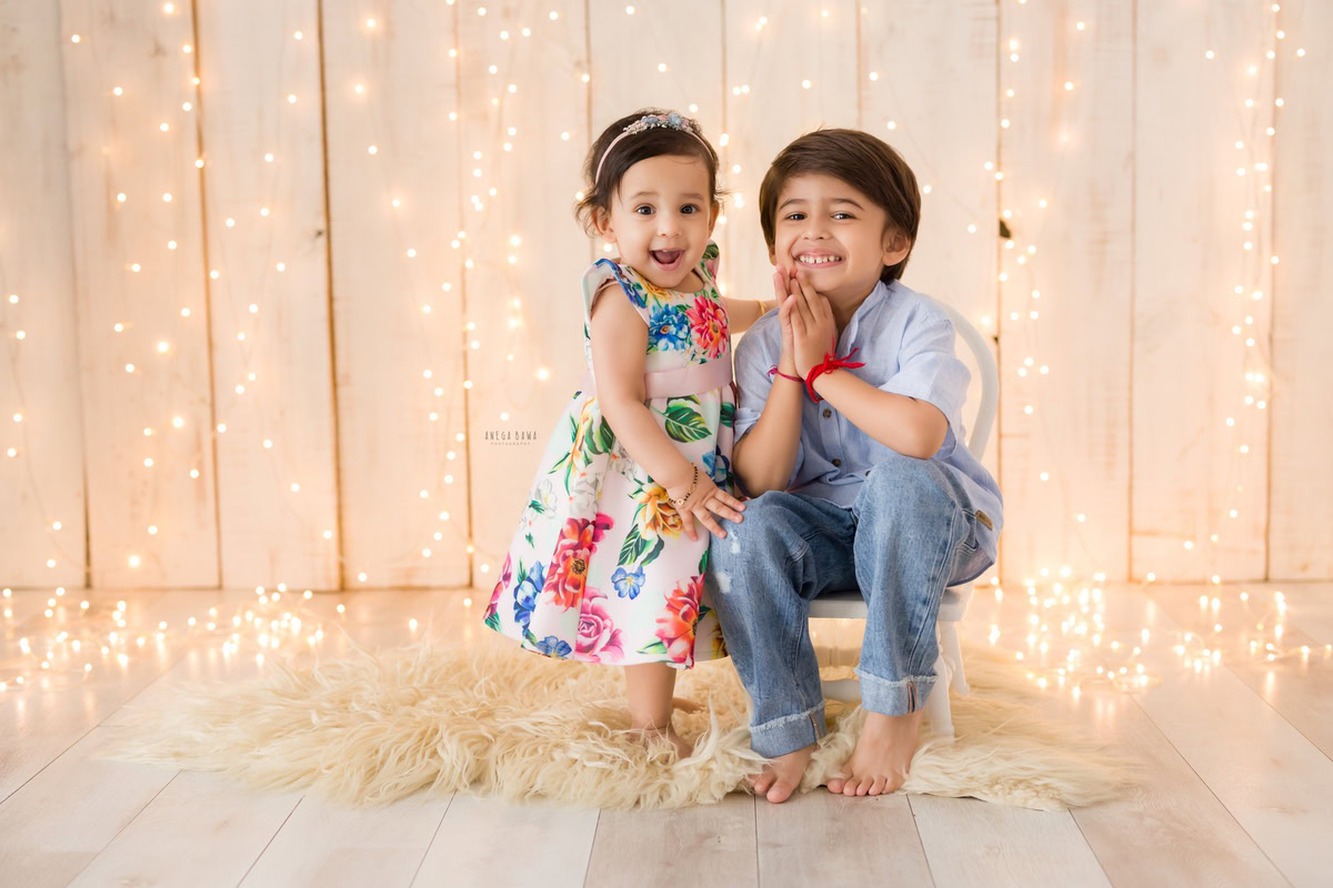 In a heartwarming moment of sibling affection, a 14-month-old girl shares a tender bond with her sibling, seated together on a cozy beige rug against a soothing beige backdrop adorned with fairy lights. This delightful scene is captured in a baby pre-birthday photoshoot in Delhi, Anega Bawa, Gurgaon, and Noida.