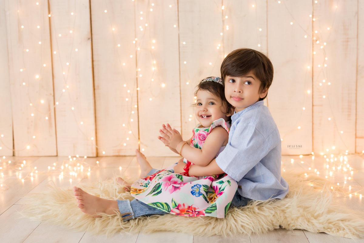 During a baby pre-birthday photoshoot in Delhi, Anega Bawa, Gurgaon, and Noida, two siblings, aged 14 months and 1 year old, share a precious moment together. They sit on a soft beige rug, illuminated by twinkling fairy lights against a serene beige backdrop, capturing the warmth of their bond.