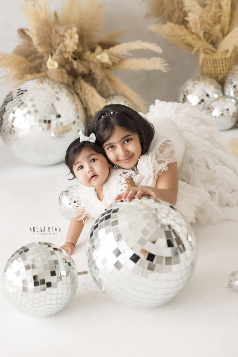 Capture the delightful bond between siblings with Anega Bawa Photography in Delhi and Gurgaon. Your 14-month to 1-year-old girl, accompanied by her sibling, poses against a backdrop adorned with shimmering discoballs and lush bushes, creating a whimsical atmosphere. Book your session now to cherish these heartwarming moments forever.