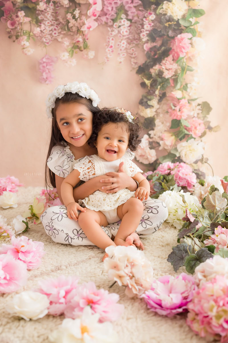 Girl with her sibling amidst a charming floral fringe, surrounded by flowers on the floor against a delightful peach backdrop, marking their journey from 14 months to 1 year old during their first birthday photoshoot in Delhi by Anega Bawa, Gurgaon, Noida.