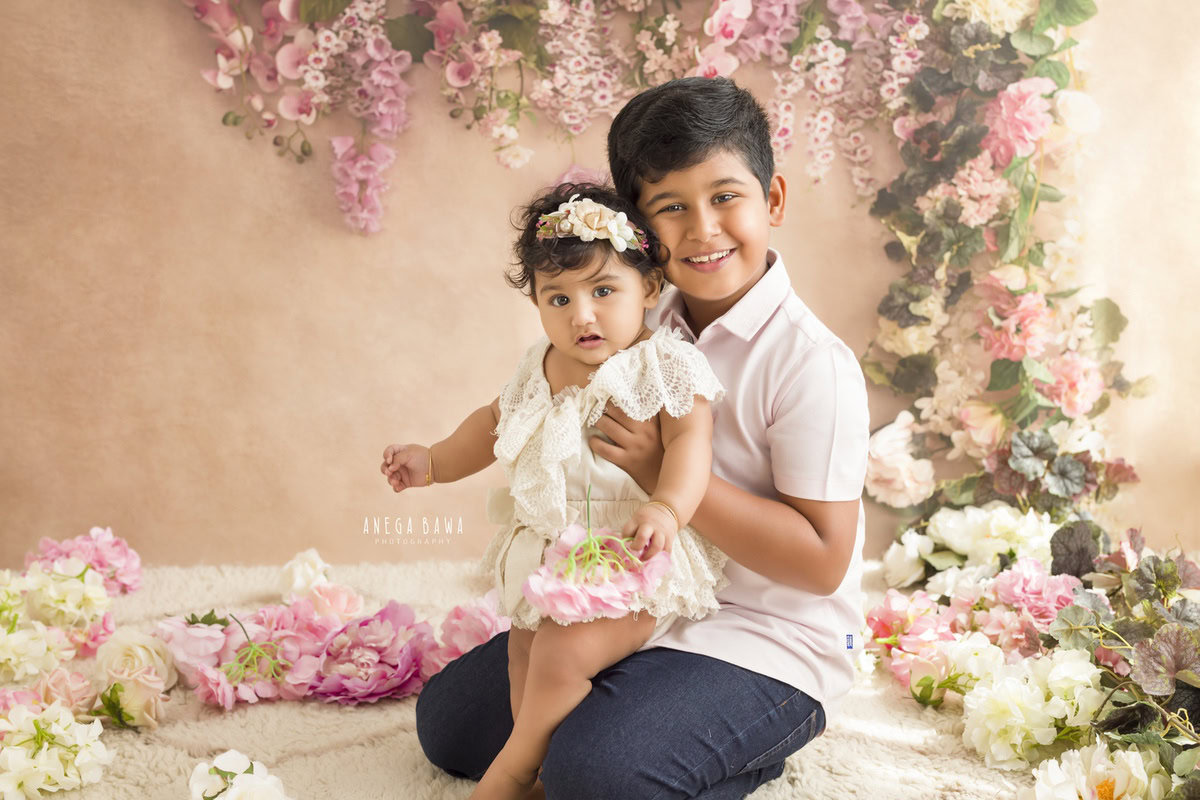 In this delightful toddler photoshoot by Anega Bawa in Delhi, Gurgaon, and Noida, two adorable siblings, aged 14 months and 1 year old, share a precious moment against a backdrop adorned with floral fringes in soft peach tones. Their endearing bond is beautifully captured, radiating warmth and innocence in every frame.