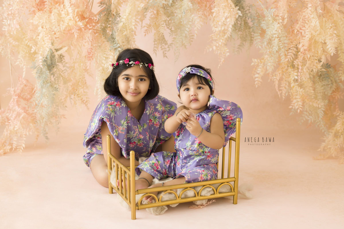 Girl with her sibling beside a regal golden cot against a backdrop adorned with leafy peachy hues, marking their journey from 14 months to 1 year old during their first birthday photoshoot in Delhi by Anega Bawa, Gurgaon, Noida.