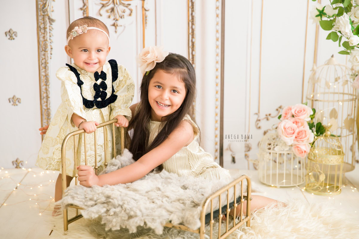 In this heartwarming shot by Anega Bawa during a sitter photo shoot in Delhi, Gurgaon, two adorable siblings, aged 14 months and 1 year old, share a precious moment. They are seated beside a golden cot, surrounded by pink flowers and castles, against a pristine white backdrop.