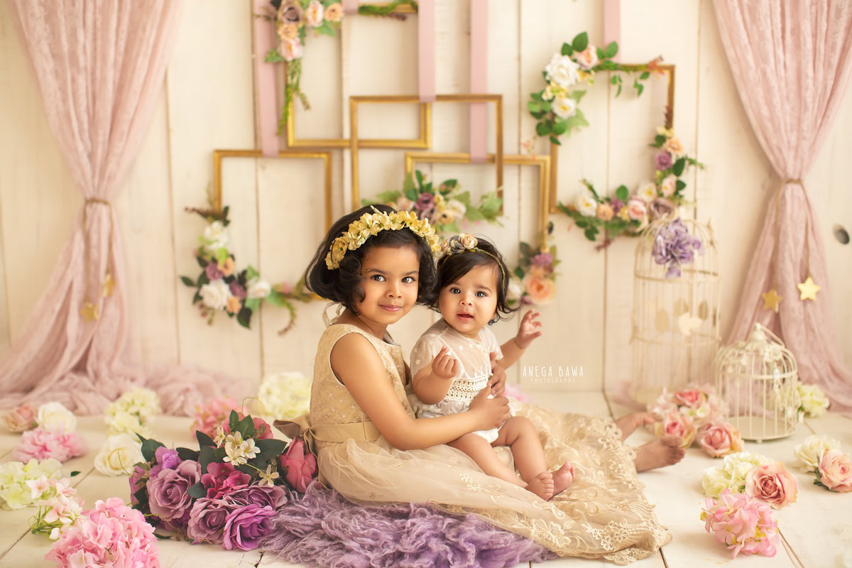 At Anega Bawa Photography's studio in Delhi and Gurgaon, your 14-month to 1-year-old princess and her sibling bask in the soft light filtering through delicate net curtains. Seated on a lavender rug against a serene beige backdrop adorned with floral frames and charming castles, they're surrounded by a sprinkling of pink and white flowers, creating a picturesque setting for capturing their sibling bond.